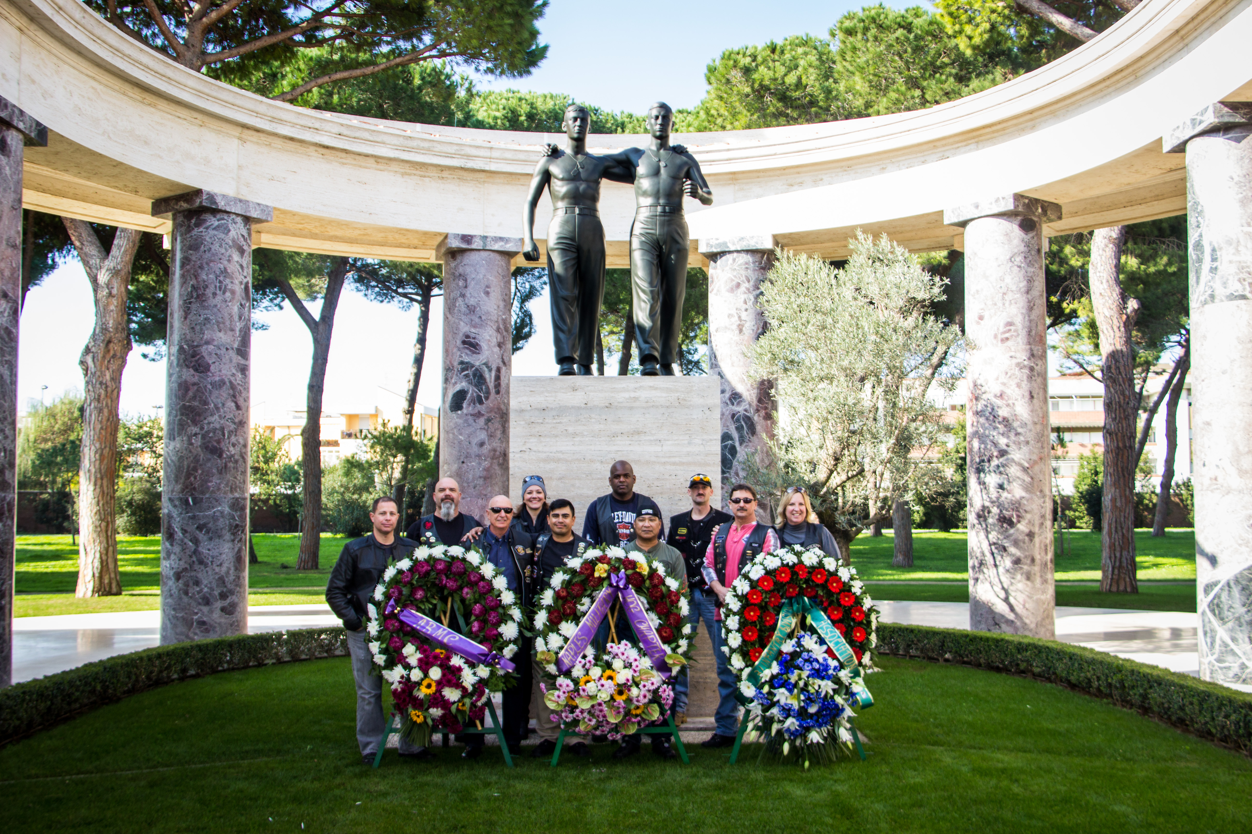 Men and women in motorcycle gear stand behind large floral wreaths after the ceremony. 