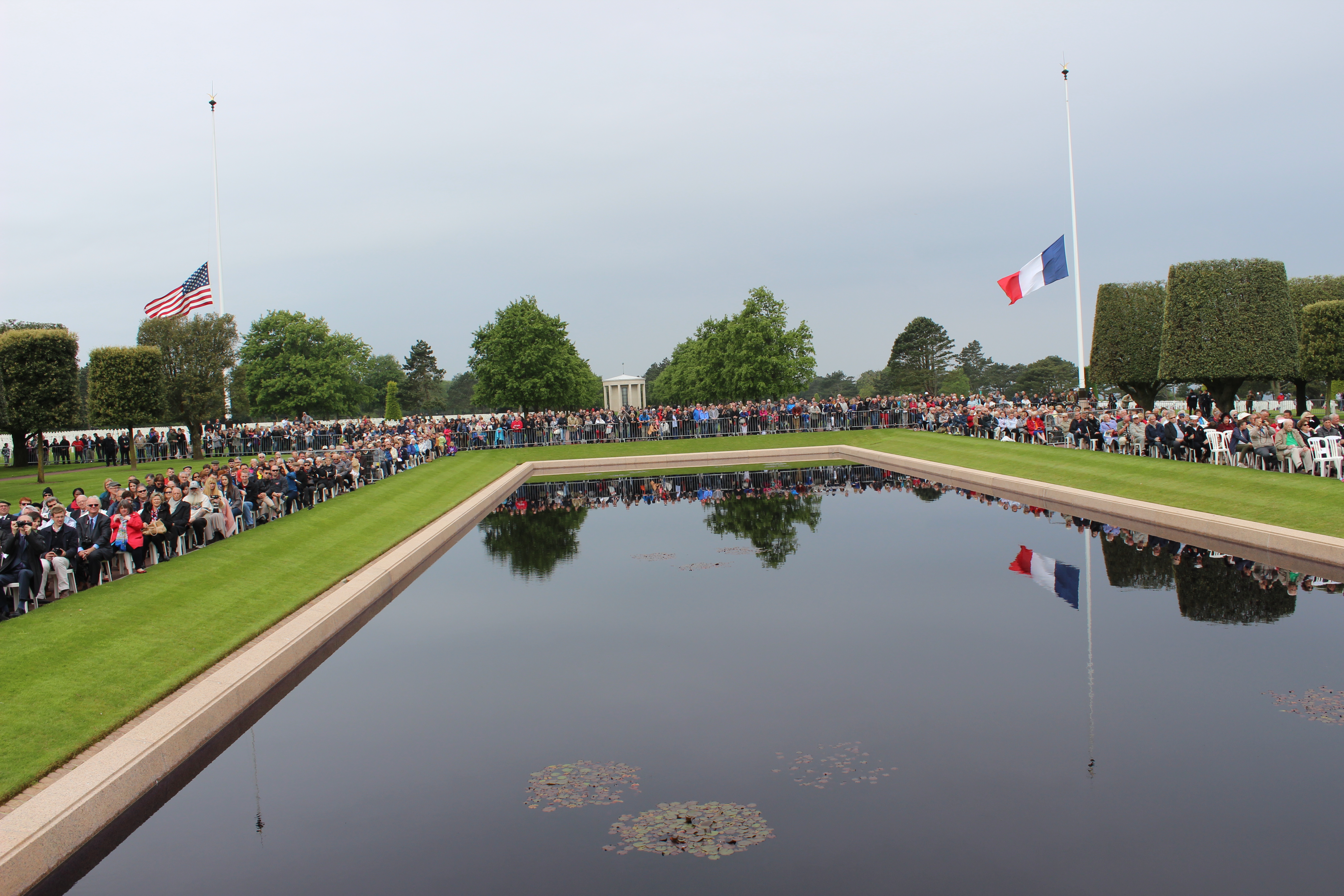 Attendees sit around the reflecting pool, with the American and French flag at half mast in the background. 
