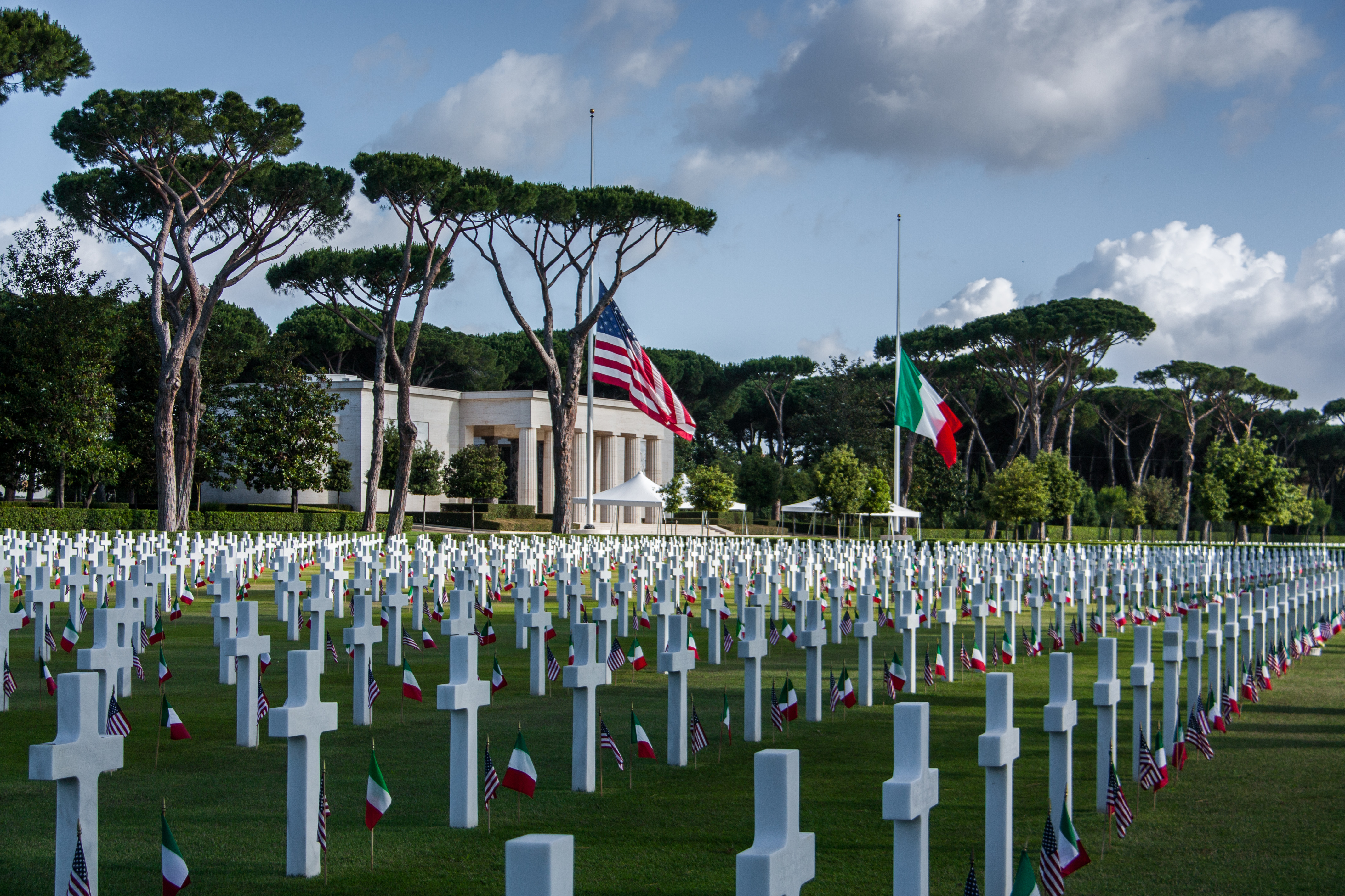 Rows of headstones with American and Italian flags cover the landscape in front of the memorial building.