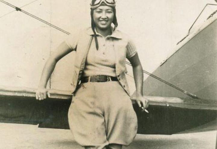 Lee, dressed in flying gear, leans against her plane.
