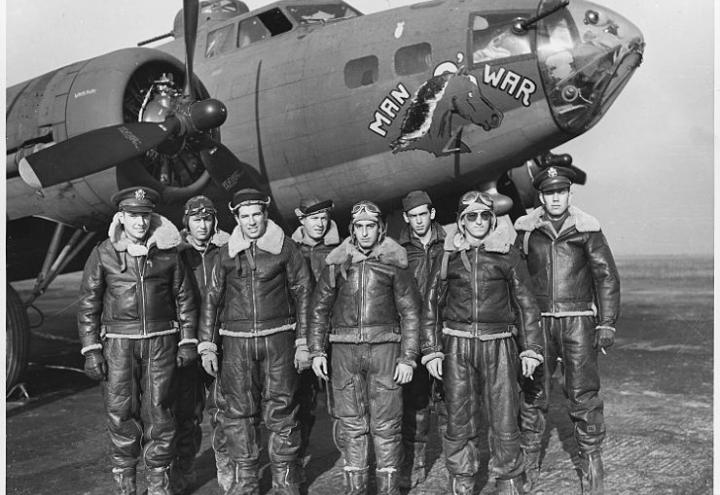 Eight men in uniform stand in front of plane in historic photo. 
