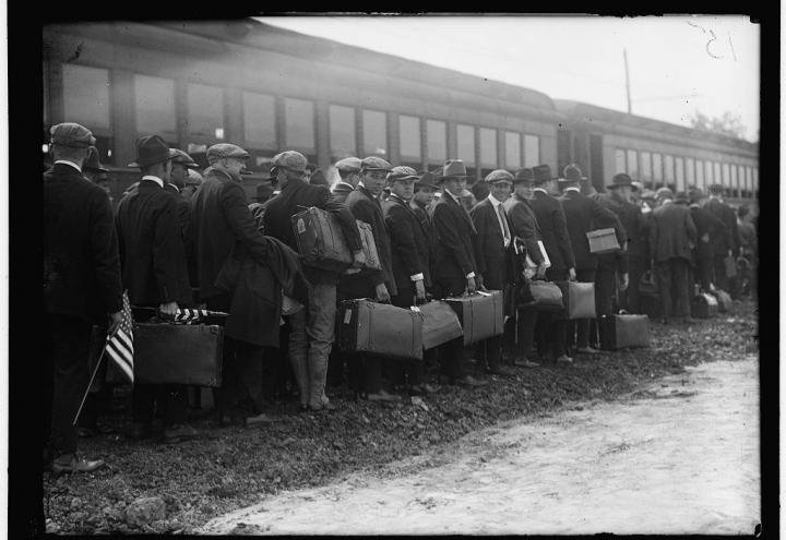 Men in suits with suitcases stand in line outside of train cars. 