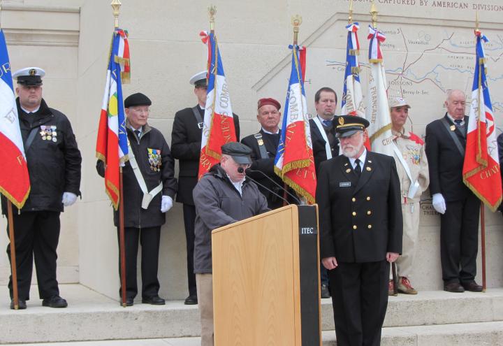 A man delivers remarks from a podium in front of a line of men holding flags.