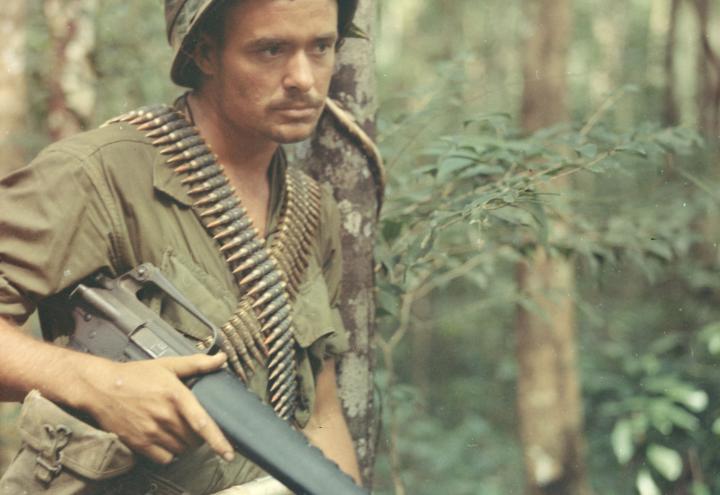 A soldier, wearing jungle-green colored fatigues, leans against a tree with weapon in hand. 