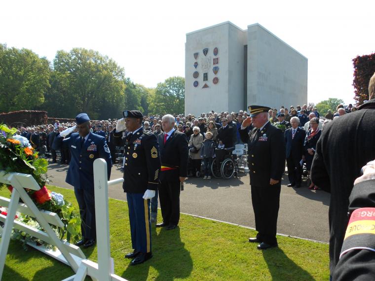 U.S. military stand saluting floral wreaths with large crowd in the background.