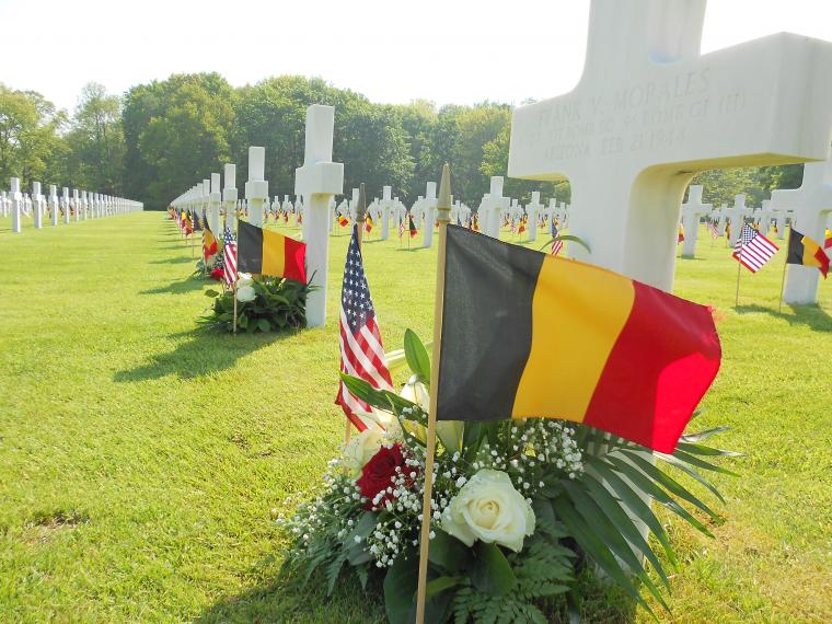 Flowers, and an American and Belgian flag rest at the base of a marble headstone.