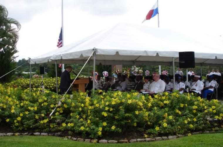 Attendees sit under tent with speaker at podium and floral bushes surround tent.