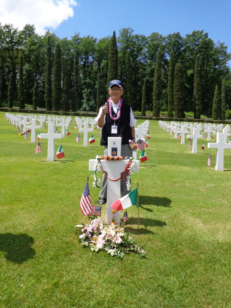 A WWII veteran stands behind a headstone that's decorated with flowers.