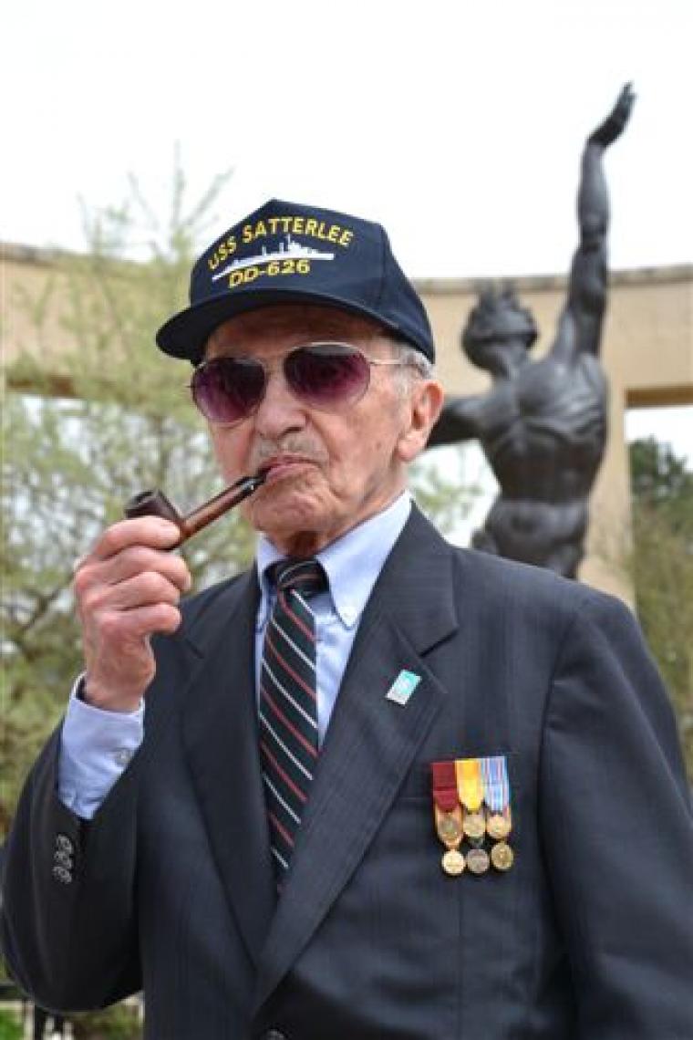 World War II veterans stand with pipe in hand, and USS Satterlee hat. 