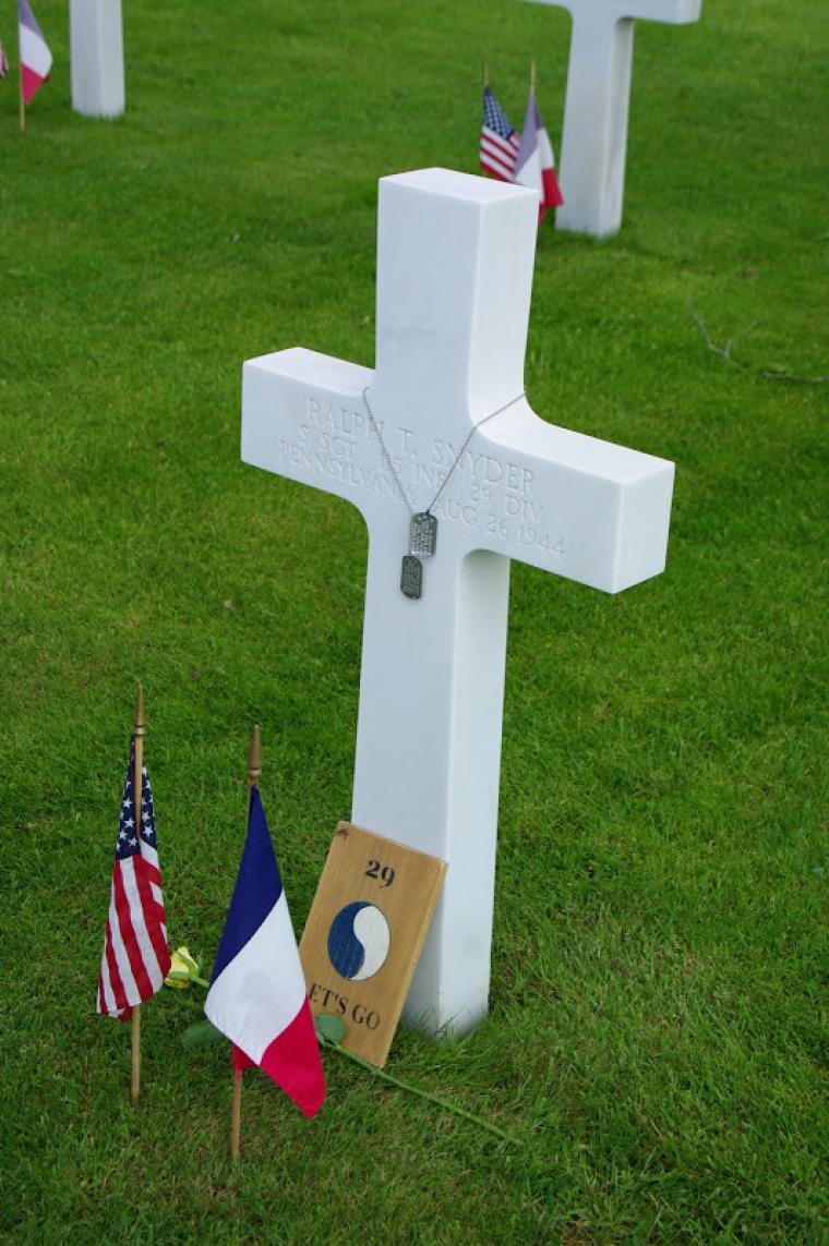 Dog tags rest on a marble headstone, while flags, a yellow rose and 29th Infantry Division sign are also present.