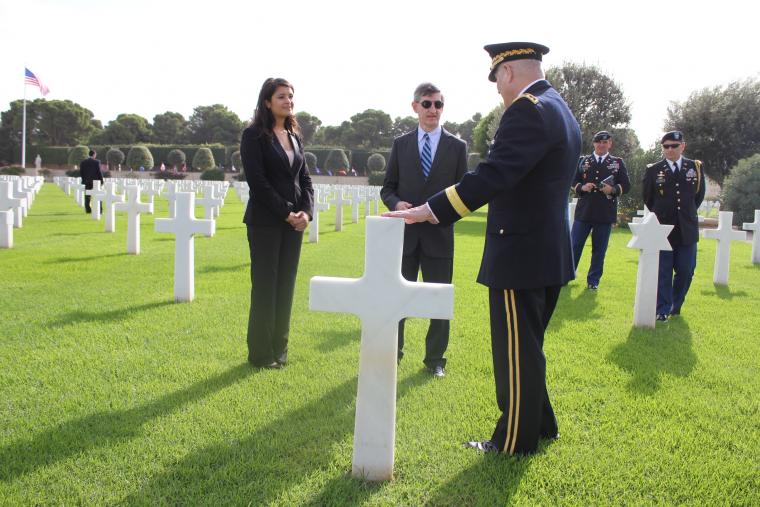 A man in uniform stands at a headstone speaking to another man and woman. 