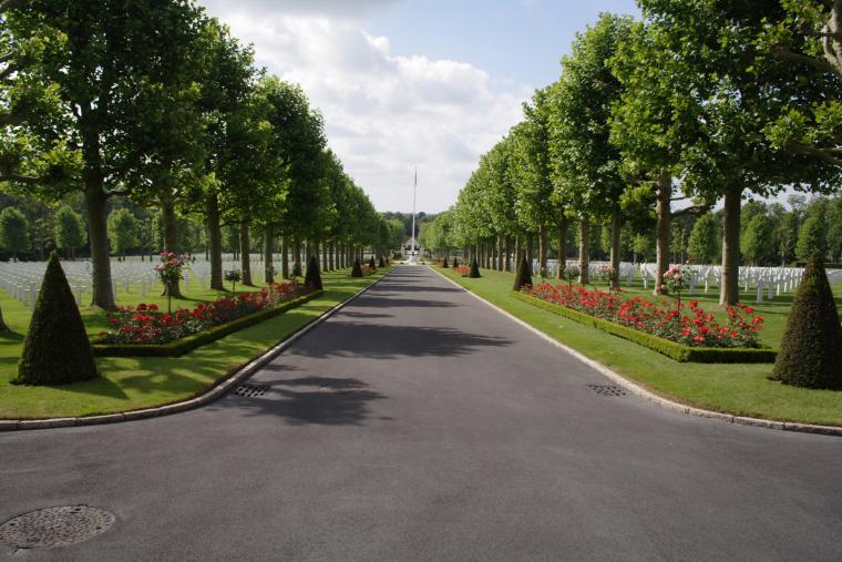 Trees and flowers line a pathway through rows of headstones at Oise-Aisne American Cemetery.