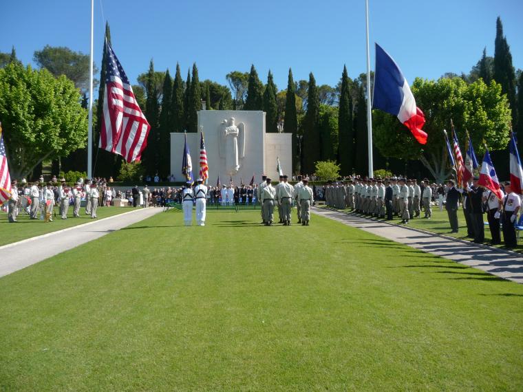 An overview of the ceremony shows participants and attendees in front of the chapel. Large American and French flags fly at half mast. 