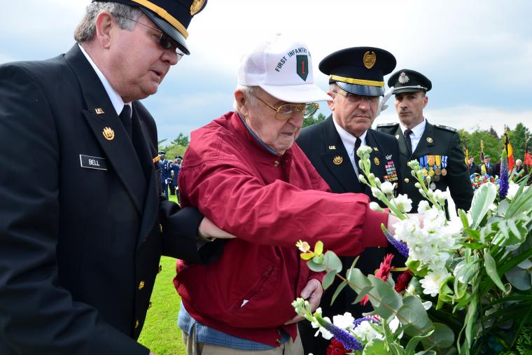 WWII veteran lays wreath, accompanied by member of U.S. military.