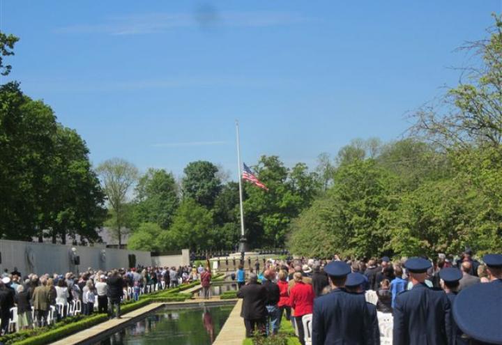 Attendees stand around reflecting pool, facing flag pole with flag at half mast.