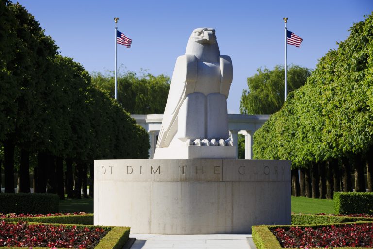 The stone American eagle sundial at St. Mihiel American Cemetery, parched atop a circular base and surrounded by Linden trees, with two American flags tin the background.