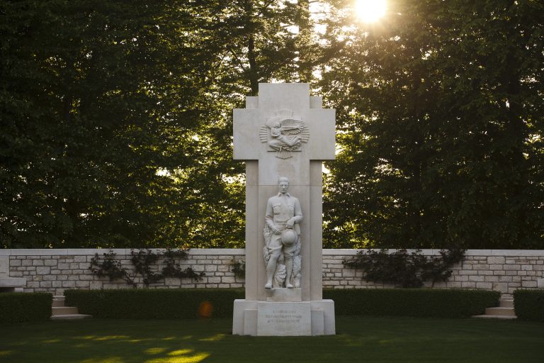 The youthful American officer monument, depicting a soldier holding a helmet stands in the sunlight, framed by lush greenery at St. Mihiel American Cemetery.