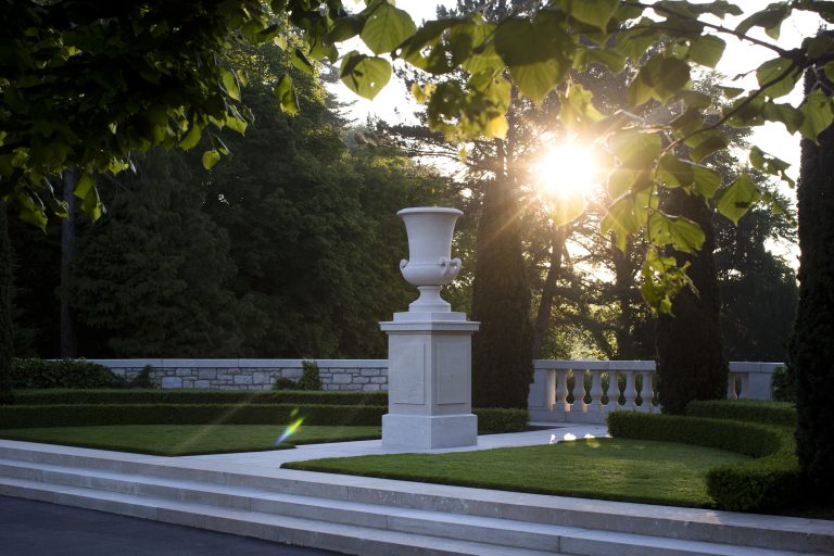 A white stone urn sits atop a pedestal with sunlight filtering through trees at St. Mihiel American Cemetery.