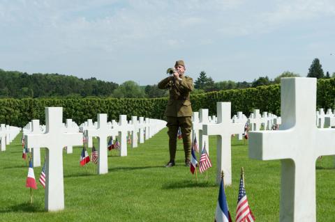 A man in a doughboy uniform plays the bugle amidst the headstones.