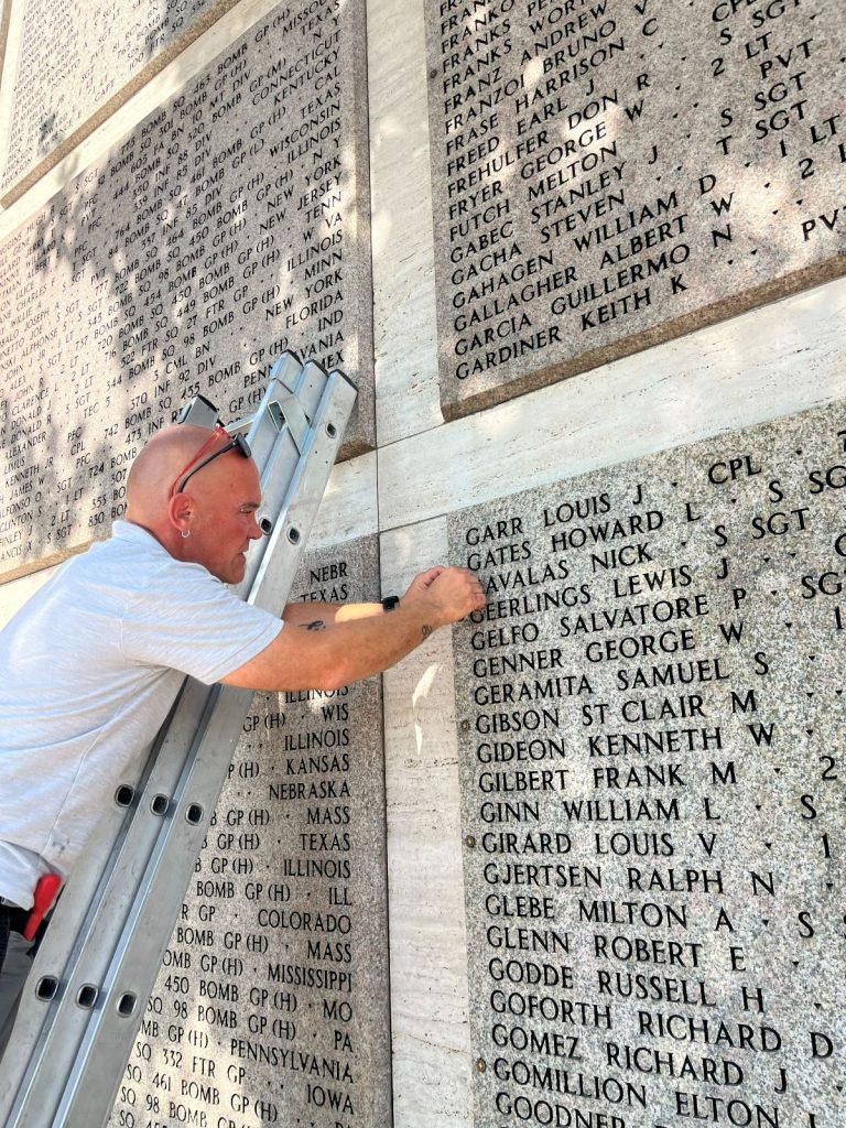A staff member at Florence American Cemetery stands on a ladder and reaches out to add a bronze rosette next to the engraved name of Nick Gavalas on the Walls of the Missing.
