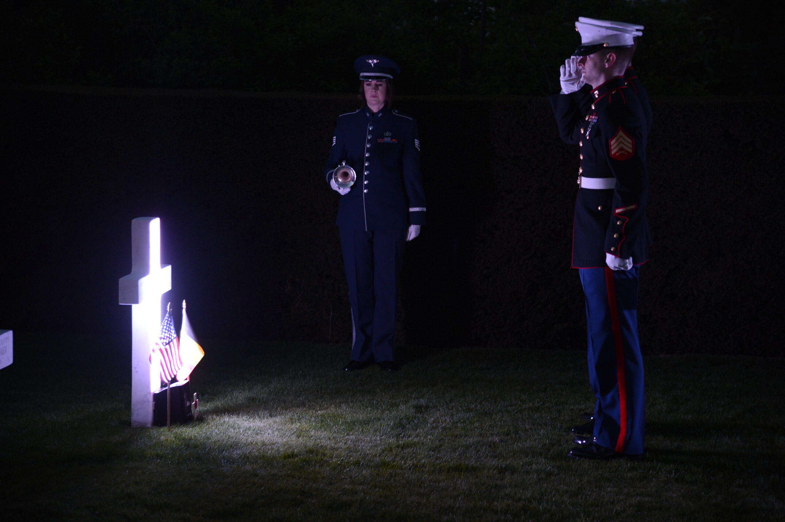 Two marines stand and salute while facing a headstone at Flanders Field American Cemetery. An Air Force bugler is also present. Image courtesy of the U.S. Embassy in Belgium.
