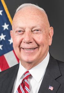 Portrait of a smiliing man with white hair in a dark suit with a red striped tie in front of an American flag.