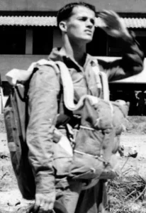 A black and white photo of Pfc. Joeseph Edgerly standing in front of a building in his flight suit and parachute saluting.