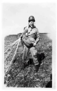 A WWII era black and white photo of Pvt. Elmer Anderson standing in a field with his parachute trailing off to his right.