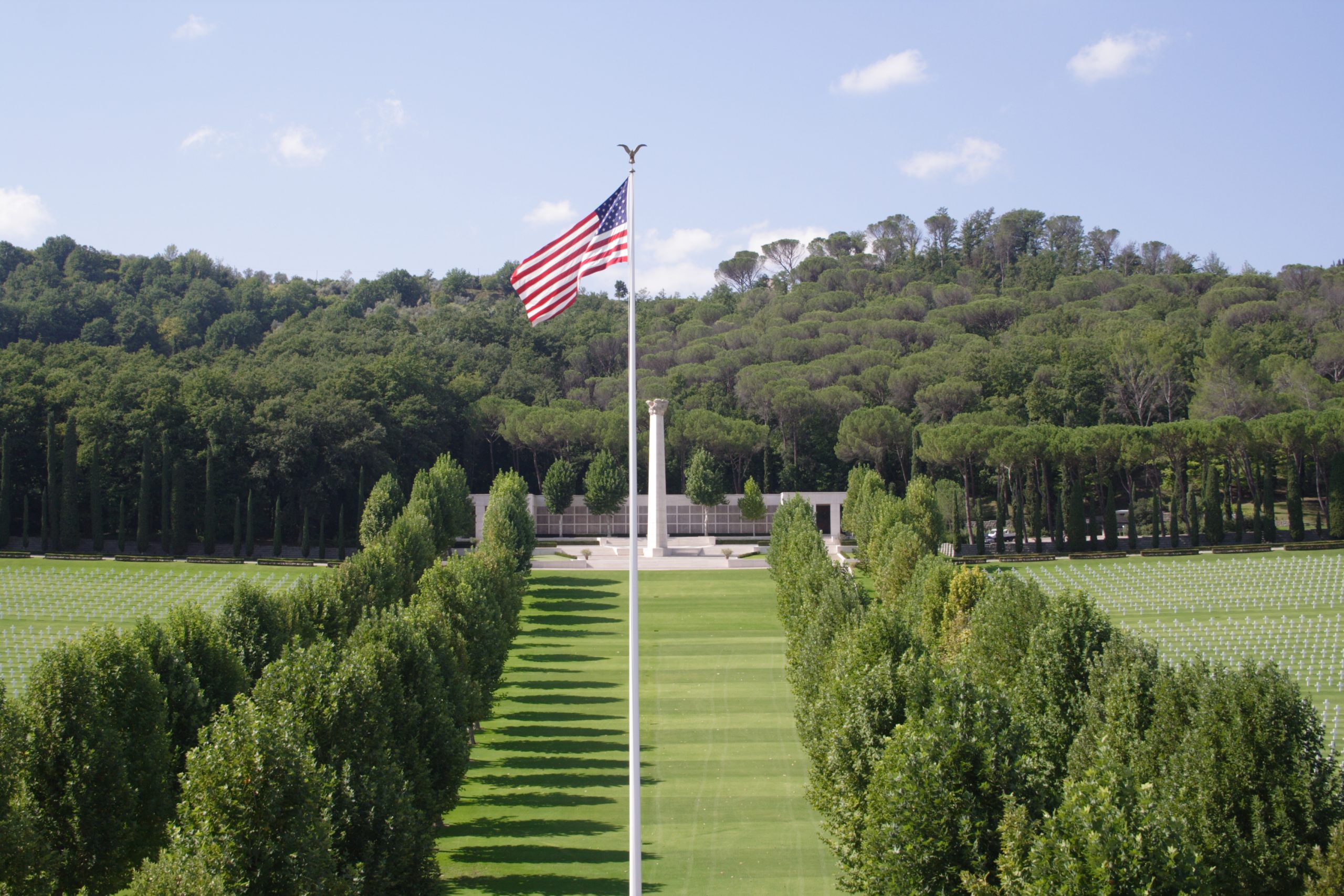 View from above of the Florence American Cemetery on a sunny day with a few clouds in the sky. The flagpole is at the foreground and the memorial in the back.