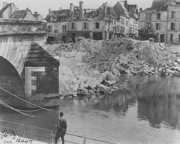 Black-and-white historic photograph of a partially destroyed stone bridge spanning a river in Château-Thierry. Large piles of rubble and collapsed masonry fill the riverbank and extend into the water near the bridge abutment. Damaged multi-story buildings with broken roofs and shattered windows line the background. In the foreground, a person stands on the river’s edge looking toward the destruction. A water-level gauge marked in meters is visible on the bridge support, documenting the extensive wartime damage to the bridge and surrounding town.