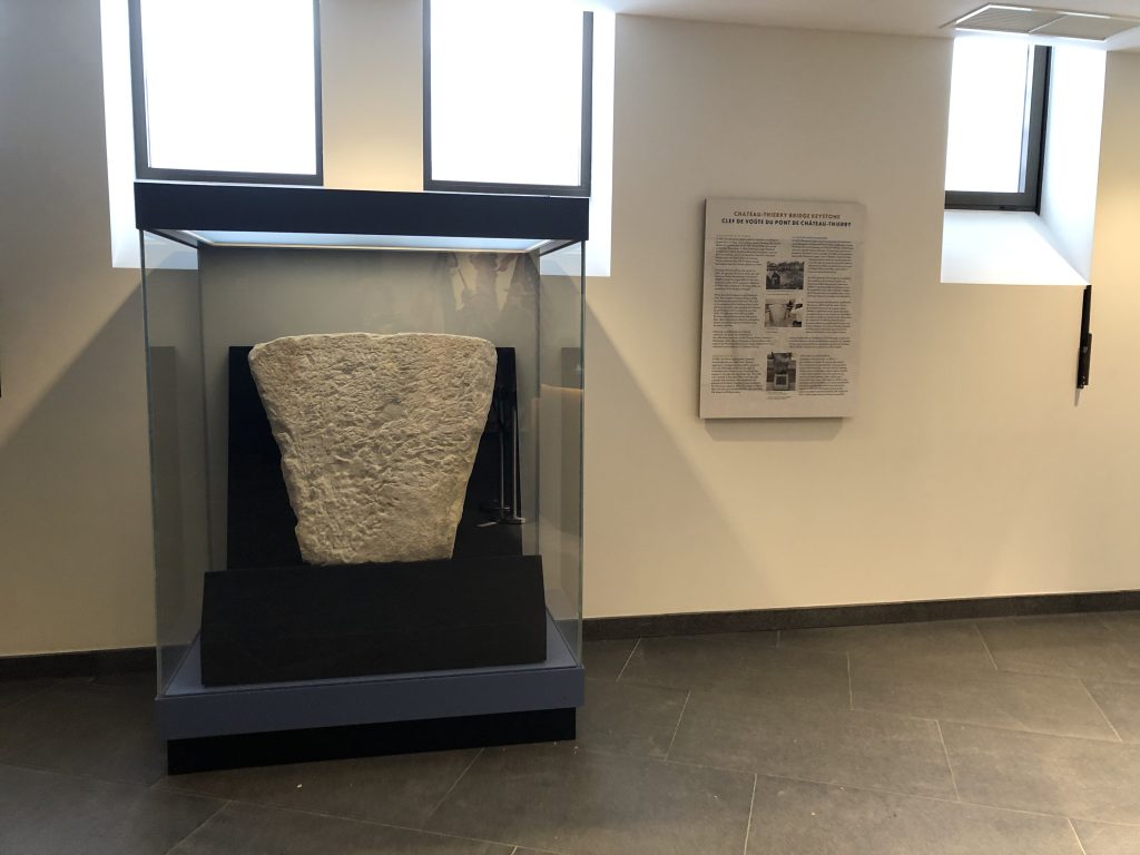 The Château-Thierry Bridge keystone is displayed inside a glass exhibit case at the Chateau-Thierry Monument. An interpretive panel nearby explains the artifact’s history, recovery, and preservation.