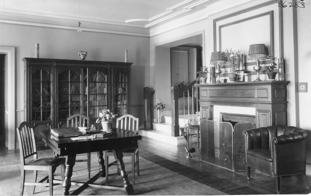 Historic black-and-white photograph of a library-style reception room with a large bookcase, wooden table and chairs, fireplace mantel, and leather armchair.