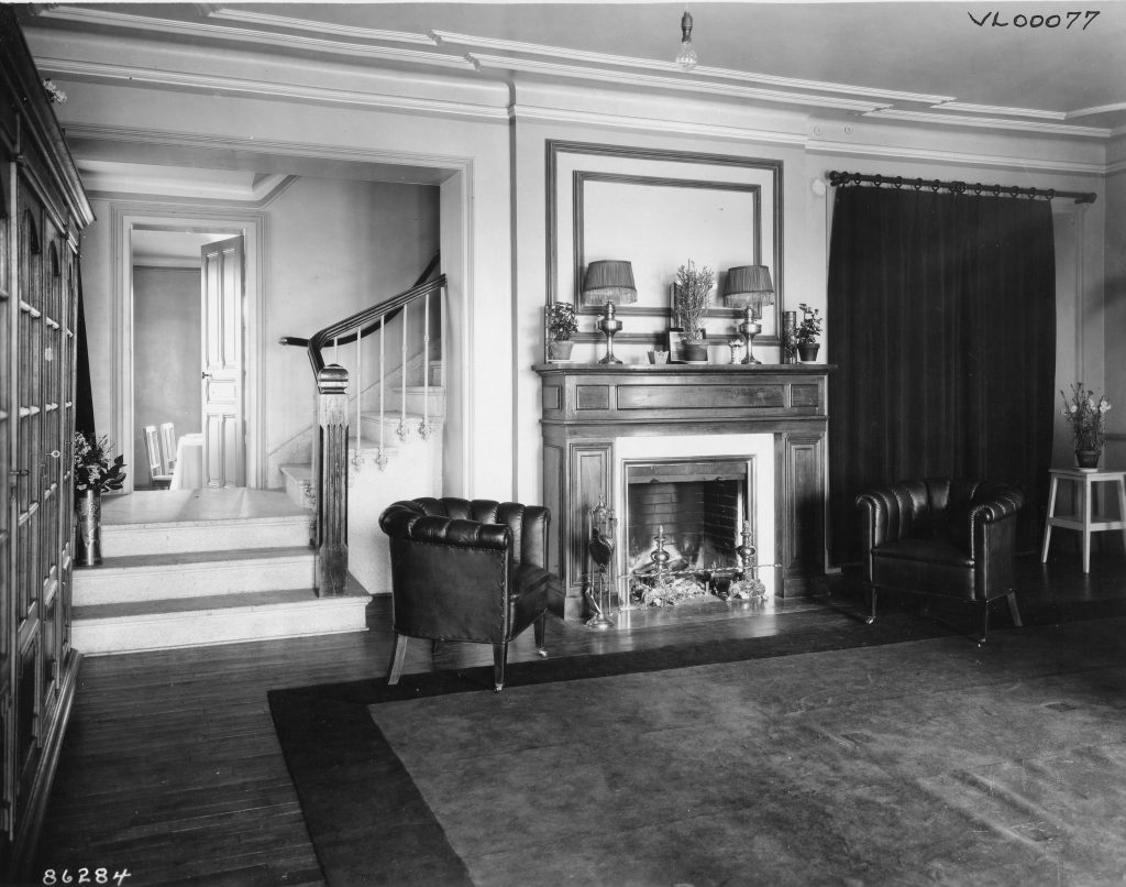 Historic black-and-white photograph of a room with a fireplace, two leather armchairs, a large bookcase, and a staircase leading to an adjacent area.