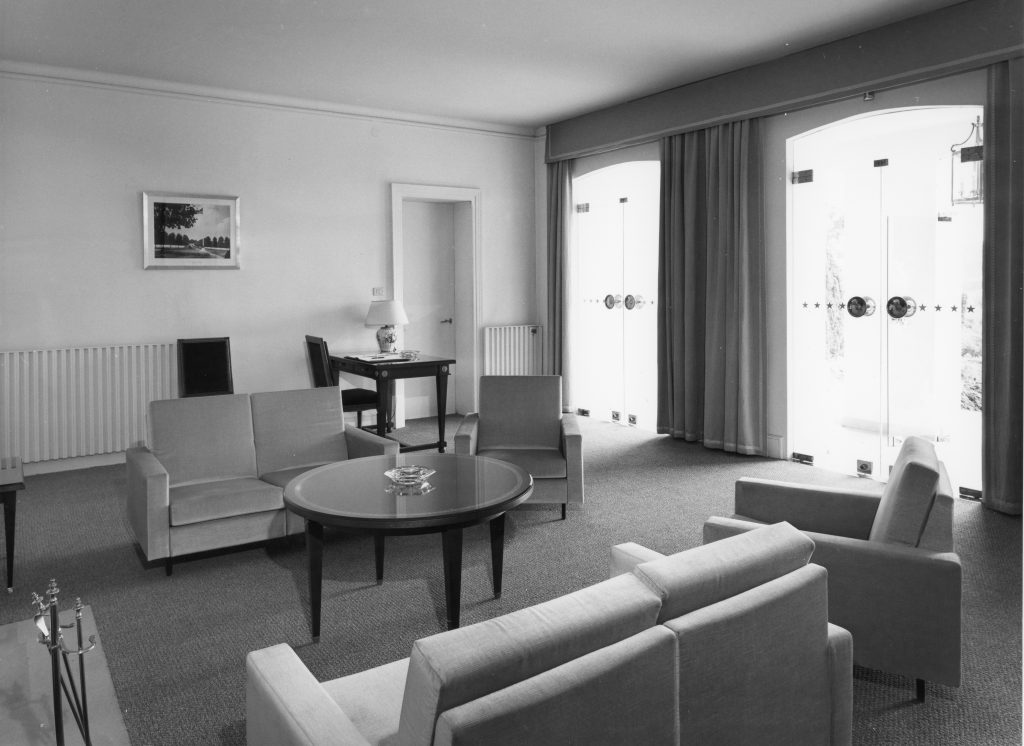 Historic black-and-white photograph of a reception room with upholstered seating arranged around a round table, a writing desk, and large glass doors letting in natural light.