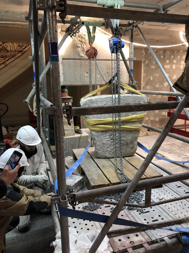 The Chateau-Thierry Bridge keystone is secured with lifting straps and chain hoists inside a scaffolded work area. Preservation specialists prepare to carefully lift and remove the historic stone for conservation and display.