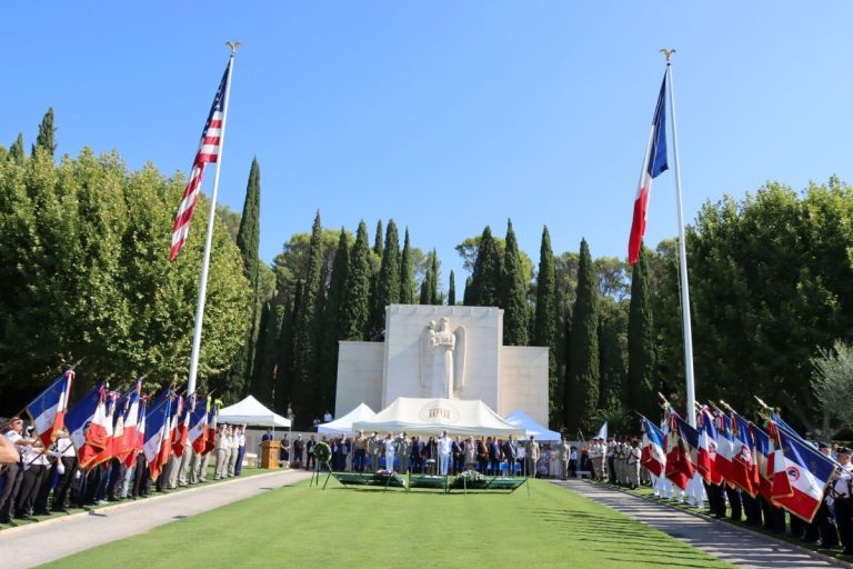A wide shot of the face of the chapel at Rhone American Cemetery with the U.S. and French flags flying on poles in front of a ceremonial tent. Racks of wreaths face the audience and flag bearers line the walkways leading to the chapel.