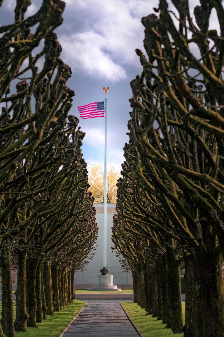 A path lined with pruned Linden trees leads to the flagpole displaying the American flag against a cloudy sky, at St. Mihiel American Cemetery.