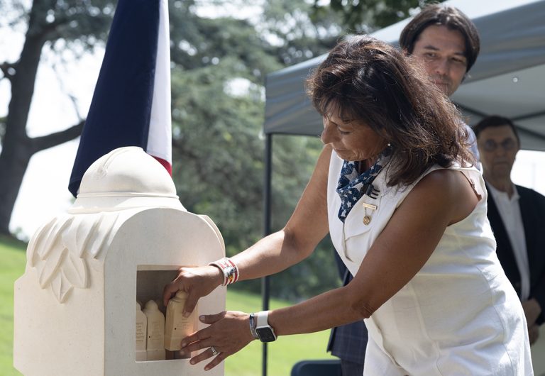 A woman places a small Sacred Soil marker into a larger white Sacred Soil marker during a ceremony at a military cemetery. A French flag stands beside her, while attendees watch from beneath a canopy. Rows of white headstones and trees are visible in the background.