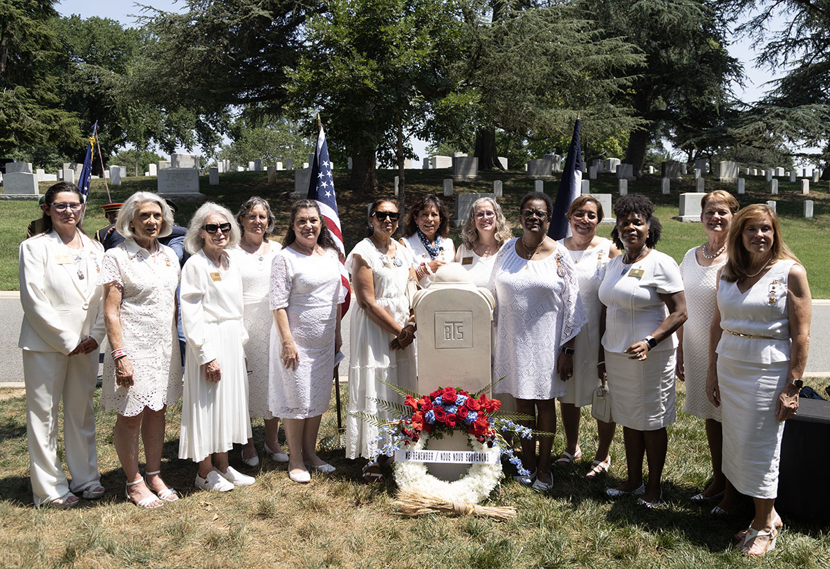 Group of American Gold Star Mothers gather around the restored Sacred Soil Marker at Arlington National Cemetery following its rededication ceremony. The women, dressed in white, standed beside the stone marker with American and French flags visible in the background. A red, white, and blue floral wreath and symbolic wheat are placed at the base of the marker.