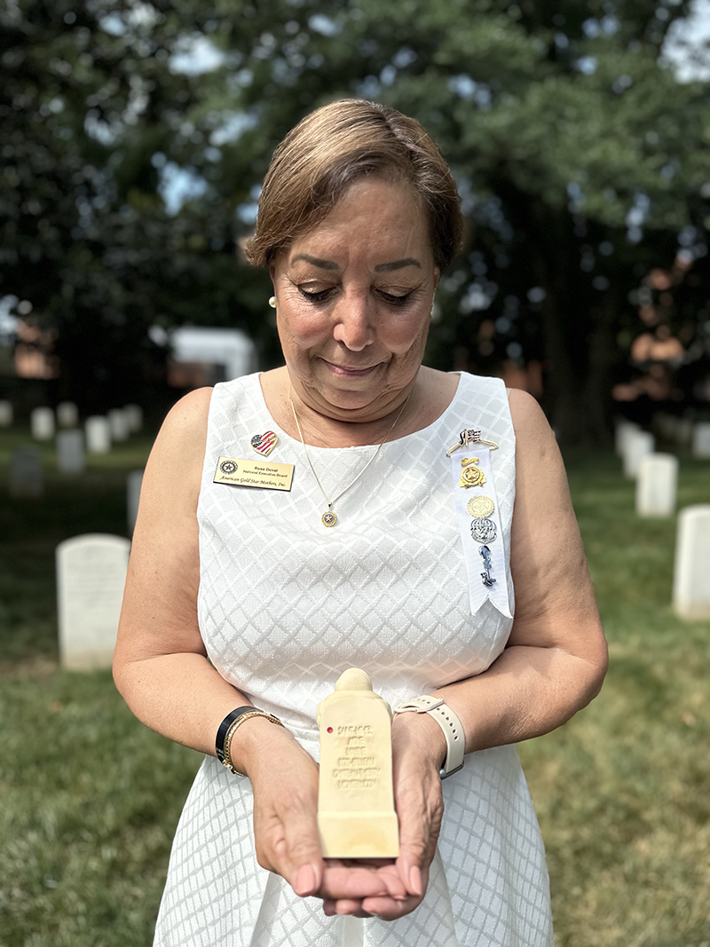 A woman stands in a military cemetery, looking down as she holds a small Sacred Soil marker in both hands. She wears a white dress and several commemorative pins, while headstones and trees are blurred in the background.