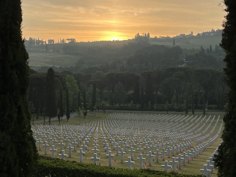 A military cemetery at sunrise with rows of white crosses. Trees line the viewing area and the sun rises over the hillside.