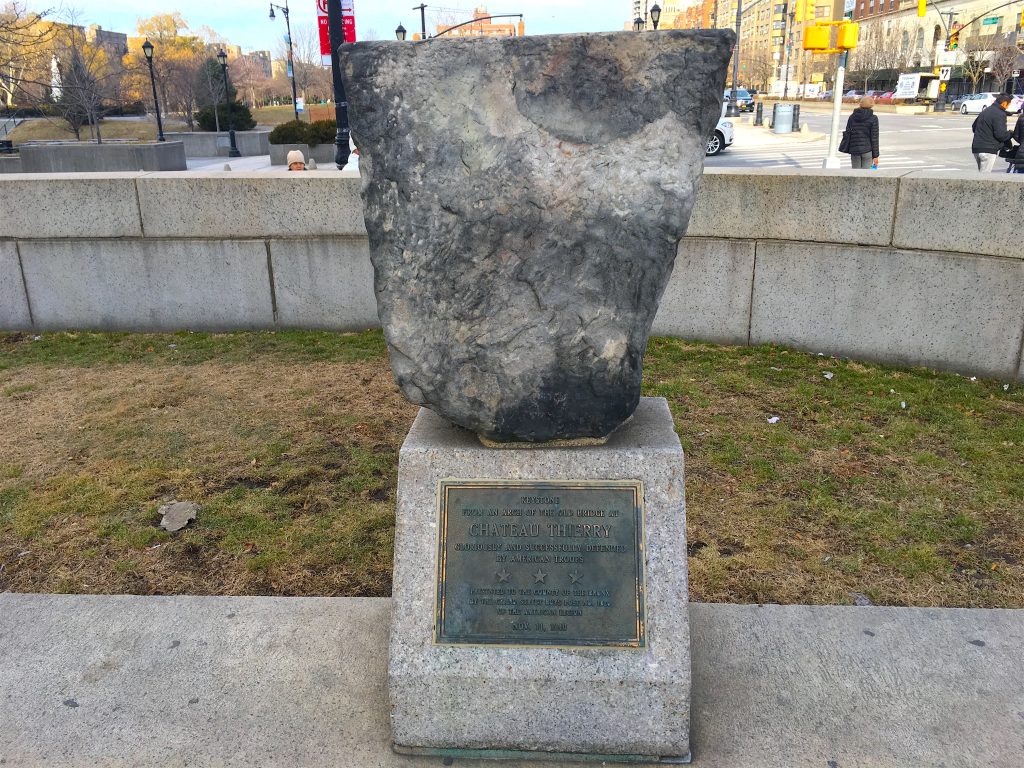 Large stone keystone displayed on a granite pedestal with a commemorative plaque honoring the defense of Chateau-Thierry by American troops during World War I. The weathered keystone stands outdoors in a landscaped urban plaza, with a low stone wall, sidewalks, streetlights, and city buildings visible in the background. The plaque identifies the artifact as a keystone from an arch of the old bridge at Chateau-Thierry.