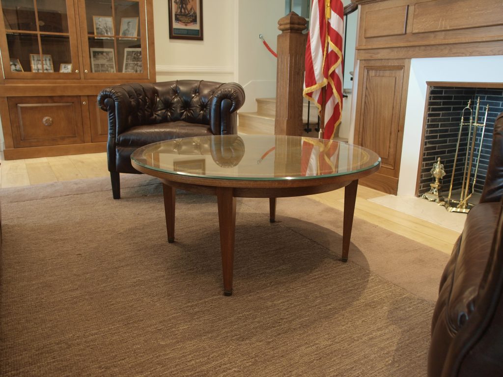 Round glass-top coffee table in a reception room with leather seating, a fireplace, and a display cabinet in the background.