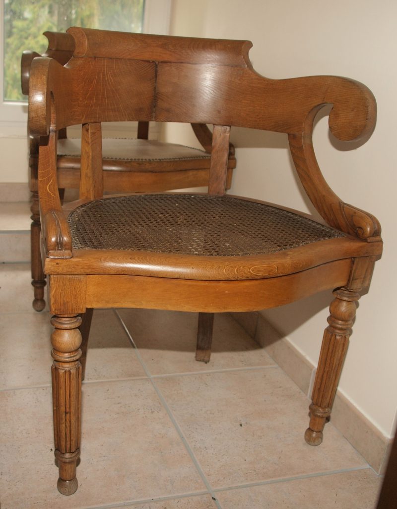 Antique wooden armchair with a woven cane seat and carved scroll arms, shown indoors on a tiled floor.