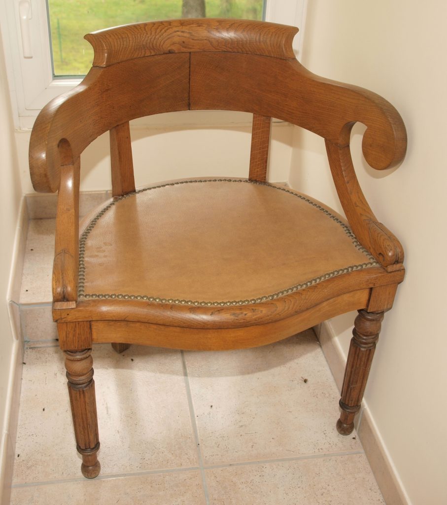 Antique wooden armchair with a leather-upholstered seat, scroll arms, and turned legs, shown indoors beside a window.